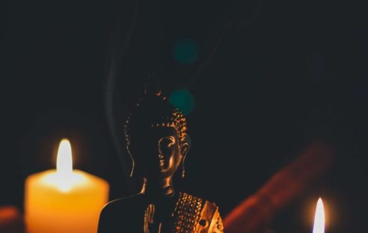 A small, dark Buddha statue in a meditative pose surrounded by two warm, lit candles in a dimmed environment.