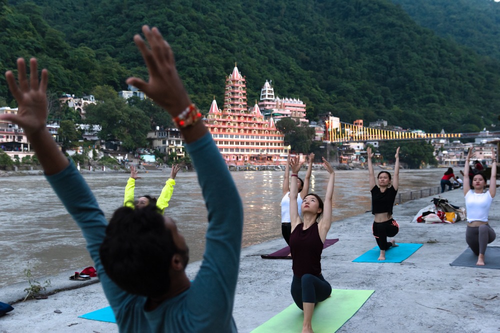 Group practicing an outdoor yoga session on mats beside a river with mountains in the background.