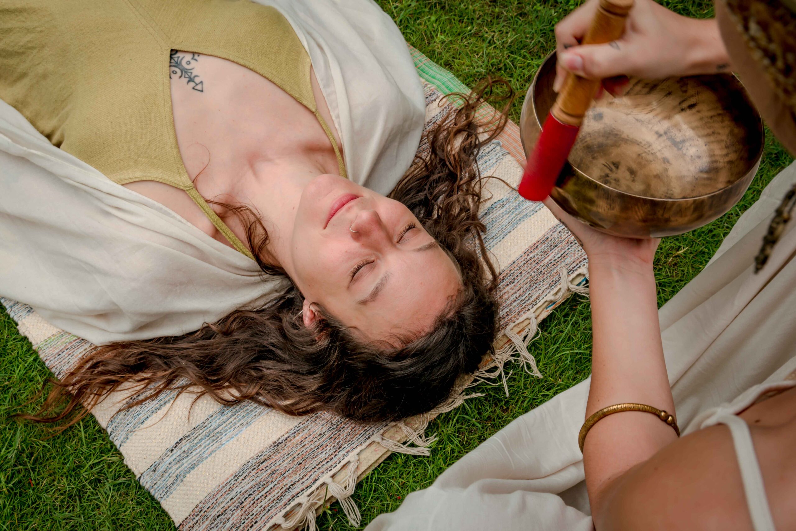 A top-down view of a woman lying peacefully on a mat outdoors, while a practitioner holds and plays a singing bowl over her.